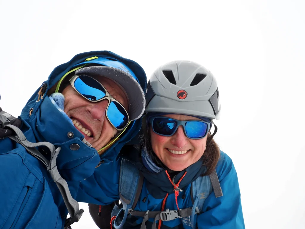 Two mountaineers in blue jackets and sunglasses taking a close-up photograph against a white background.