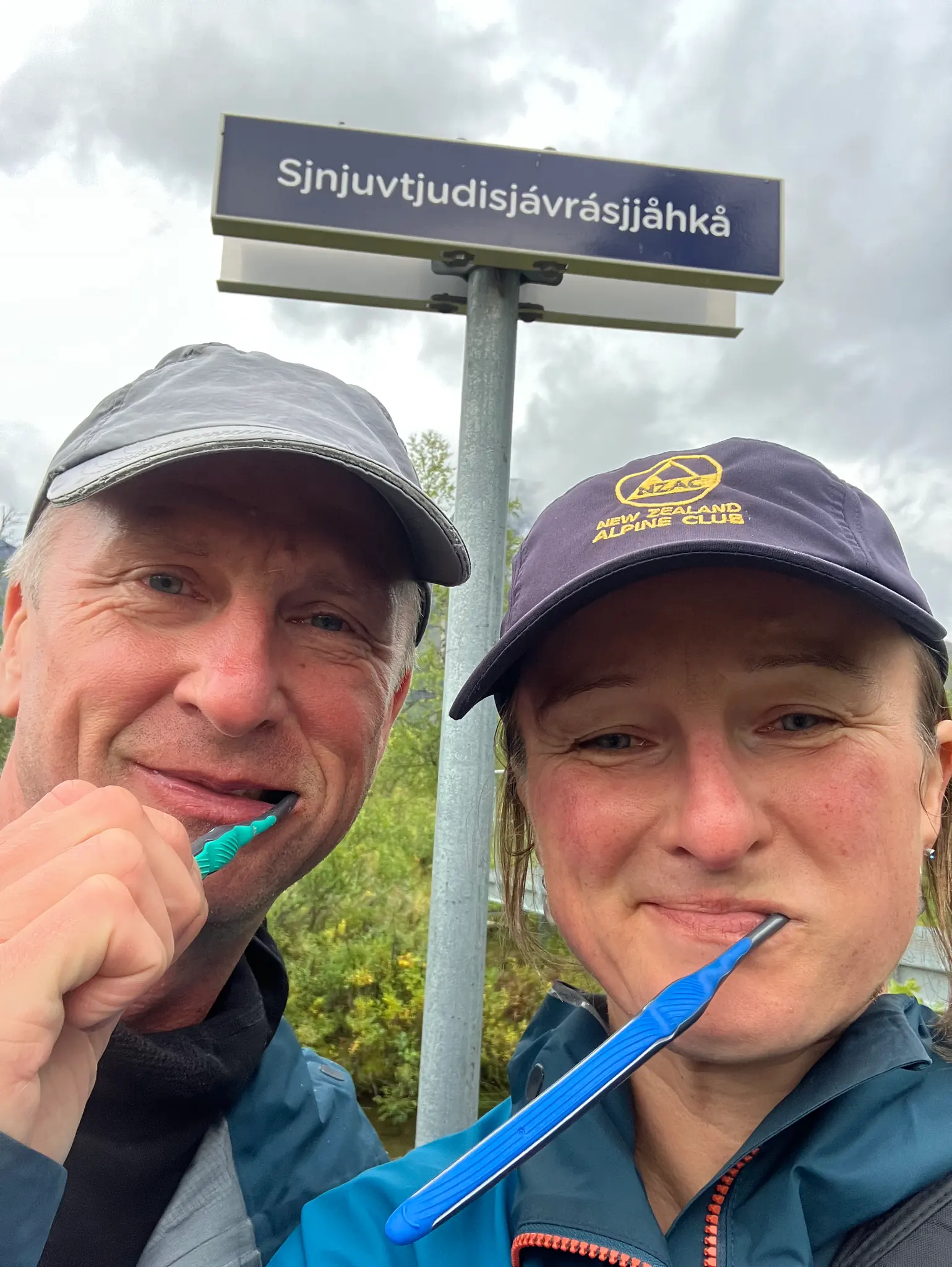 Two travellers brushing their teeth beneath a Sámi place-name sign.