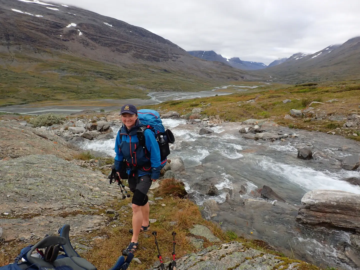 Joanna standing beside a rocky mountain stream with a broad river valley behind. Sarek NP, 2026