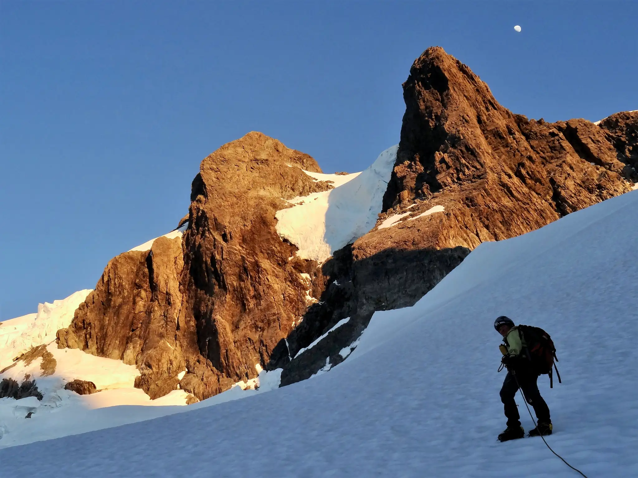 Middle section of the snowy slope on Mount Tutoko. Rock towers lit by the rising sun behind and the moon above.