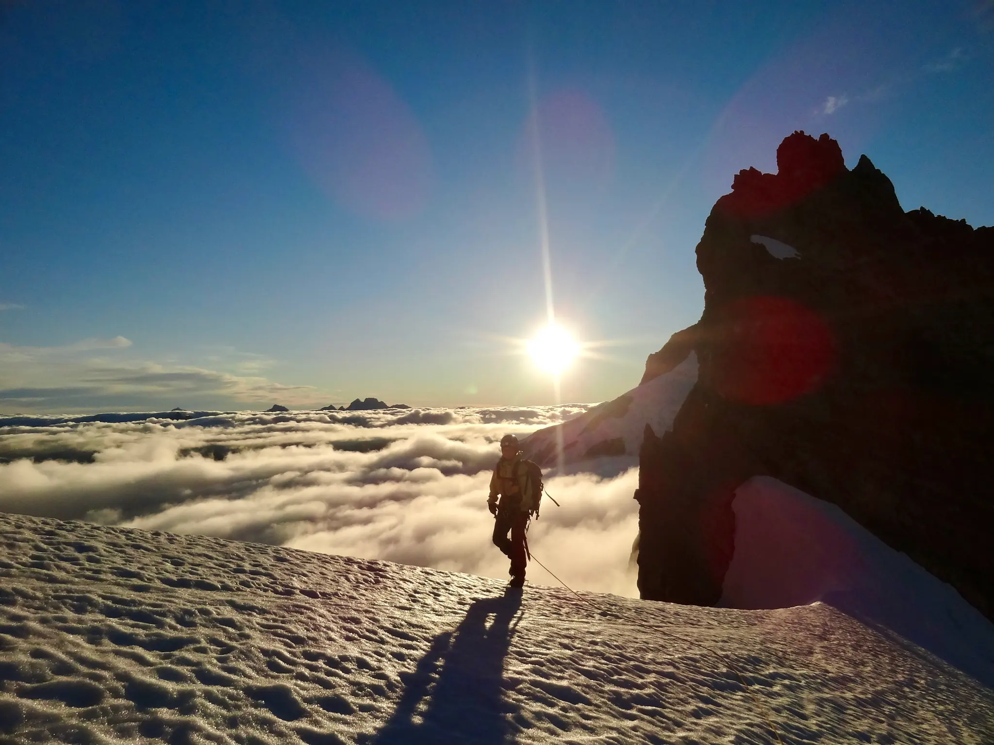Joanna at the beginning of Mount Tutoko's snowy ridge, overlooking a sea of clouds in the valley on the other side, with the sun low in the sky.