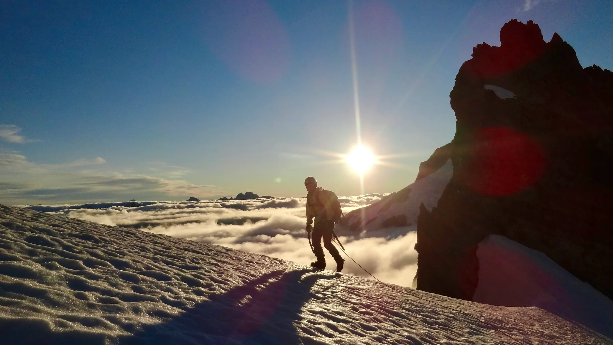 Joanna at the beginning of Mount Tutoko's snowy ridge, overlooking a sea of clouds in the valley on the other side, with the sun low in the sky.