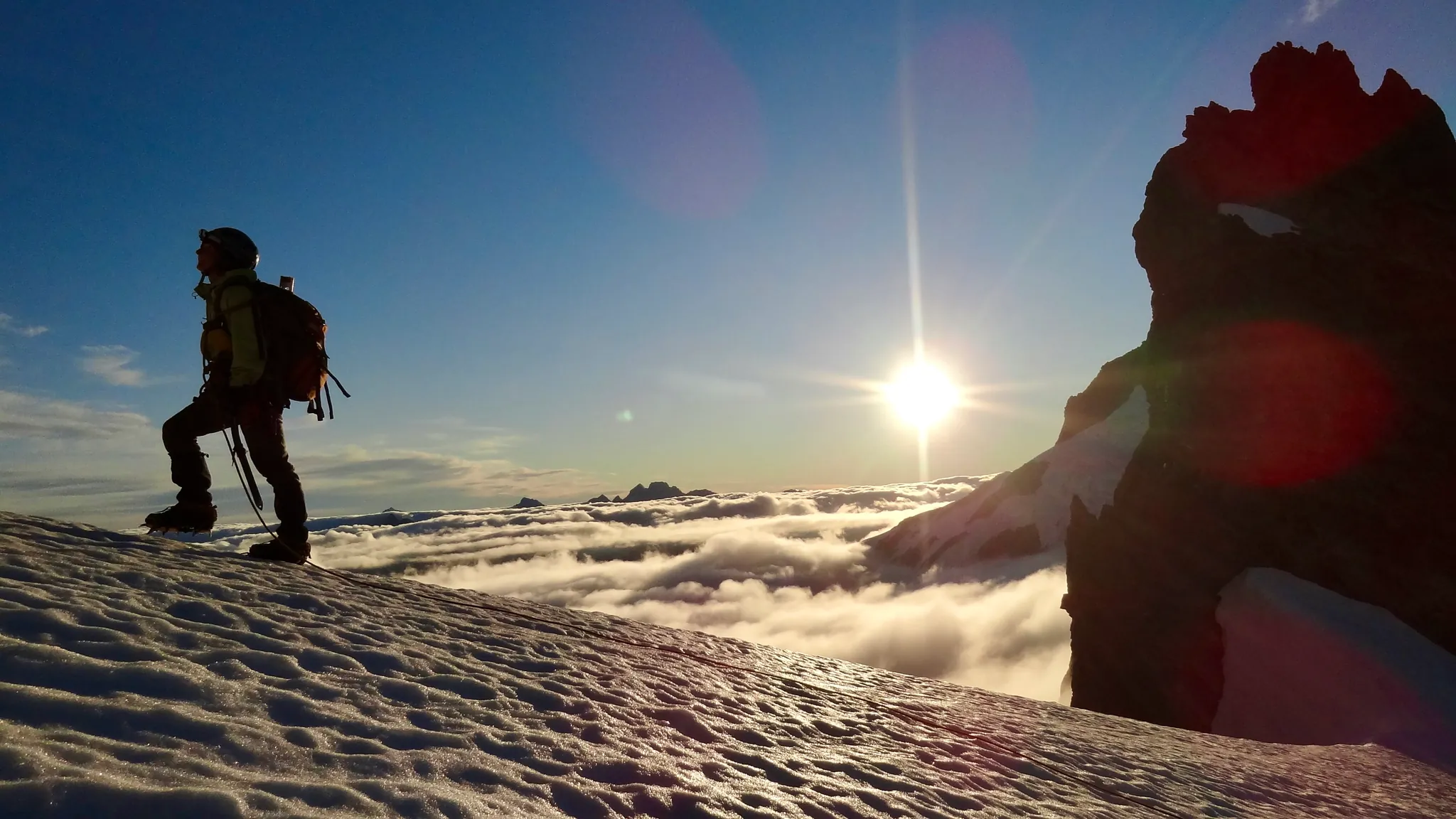 Joanna at the beginning of Mount Tutoko's snowy ridge, overlooking a sea of clouds in the valley on the other side, with the sun low in the sky.